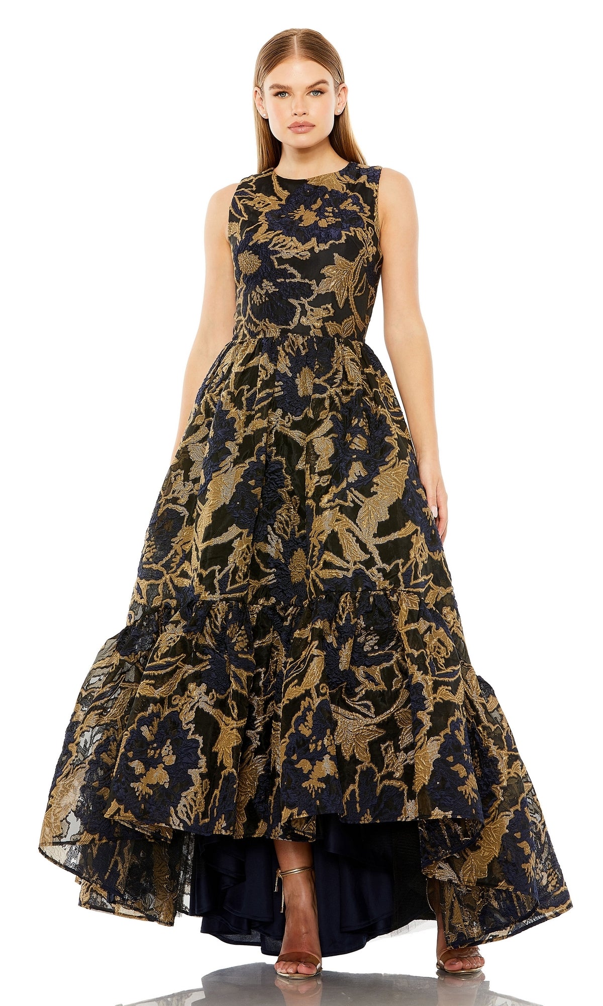 A woman wears the Mac Duggal 20714 Brocade High-Low Formal Dress, featuring a sleeveless bodice, gold and navy floral pattern, and tiered skirt. She has straight hair and completes her look with open-toed heels.
